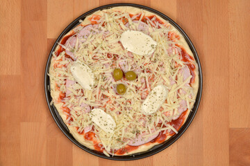 An unbaked pizza, generously topped with shredded cheese, ham, fresh mozzarella, and green olives, is ready for the oven