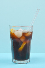 A tall glass of dark soda with ice and a lemon slice, featuring a clear straw, is set against a light blue background