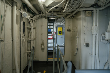 A narrow corridor inside a military ship shows exposed cables, metal pipes, and a yellow safety sign on a door in the background. The industrial space is dimly lit and utilitarian in design.