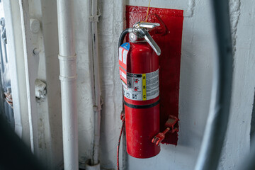 A red fire extinguisher is securely mounted on a white metal wall with a red backplate. It is...
