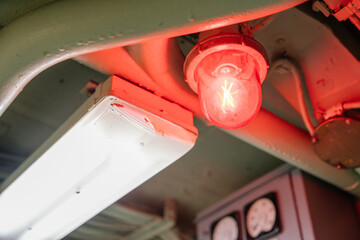 A close-up of a glowing red warning light next to a white fluorescent light on a metal ceiling. Industrial pipes and gauges are visible in the background.