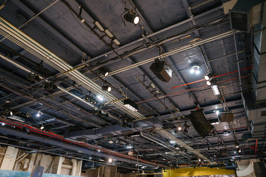 Detailed view of the ceiling inside the Intrepid Museum showing lighting equipment, pipes, and speakers. The exposed industrial infrastructure supports sound and light systems for exhibits and