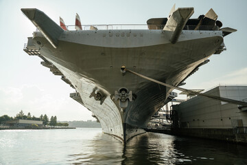 Frontal view of the USS aircraft carrier docked in New York, with the ship's bow reflecting on the calm water surface. The massive anchor and hull details are clearly visible. © DenisProduction.com
