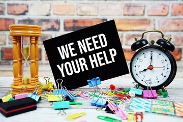 We Need Your Help text on paper card with alarm clock and stationary on wooden background