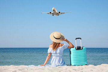 Woman with suitcase on sandy beach looking at airplane flying in sky, back view