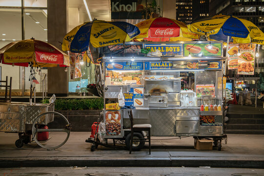New York City, New York, USA - 07.30.2024: A vibrant halal food cart is parked on a city sidewalk at night, decorated with bright umbrellas and illuminated signage. The cart displays images of hot