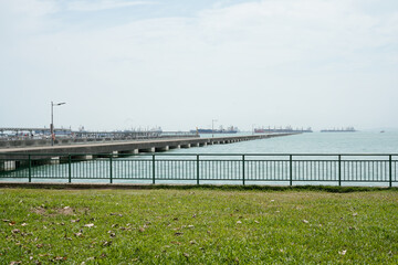 Fototapeta premium view of a pier stretching over the blue-green ocean waters under an overcast sky, with docked ships visible in the distance