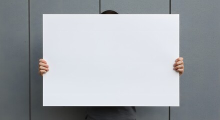 Person holding a large blank white sign in front of a gray wall.