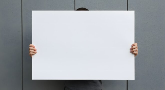 Person holding a large blank white sign in front of a gray wall.