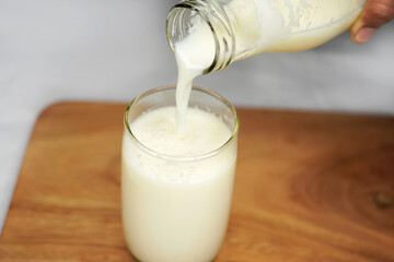 Pouring milk into the glass mug on a white table. Cow's milk from farm in jug bottle. White...