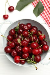 Fresh wet cherries in colander and leaves on white wooden table, flat lay
