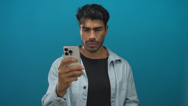 Young hispanic man holding smartphone in blue studio looking at screen with focused expression; online anonymity privacy concealment.