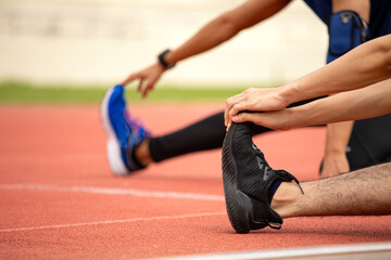 Close-up of two male athletes stretching legs on red track with focus on black running shoe and hand