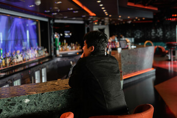 A person sits contemplatively at a bar counter with vibrant lighting and glass decor, conveying a serene and stylish atmosphere in a modern bar setting