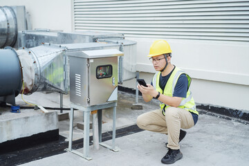 asian engineer working on smart phone with  external unit of commercial air conditioning and ventilation system installed on industrial building roof