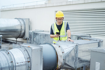 Naklejka premium asian engineer working on smart phone with external unit of commercial air conditioning and ventilation system installed on industrial building roof