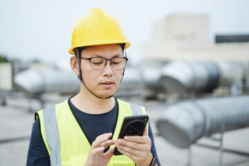 asian engineer working on smart phone with  external unit of commercial air conditioning and ventilation system installed on industrial building roof