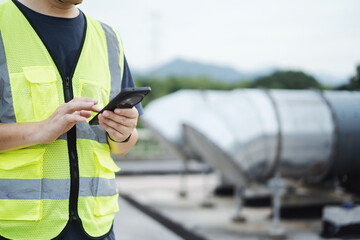 asian engineer working on smart phone with  external unit of commercial air conditioning and ventilation system installed on industrial building roof