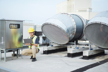 asian engineer working on laptop with  external unit of commercial air conditioning and ventilation system installed on industrial building roof