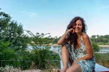 portrait of a smiling middle-aged woman in a blue summer dress sitting on a table at sunset by the...