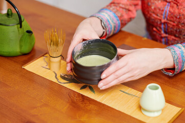 Serene tea ceremony with woman holding black teacup of matcha tea, surrounded by traditional teaware on a wooden table, capturing tranquility, heritage, and mindful ritual in a minimalist setting