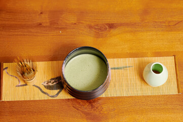 Serene tea ceremony scene with vibrant green matcha in black ceramic cup, accompanied by bamboo whisk and small white vase on a wooden table, evoking calmness and cultural tradition