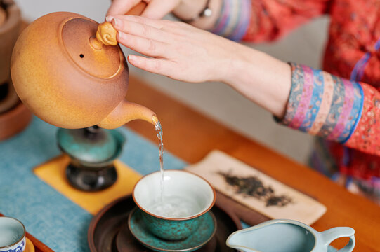 Woman pouring hot water from traditional teapot into teacup, surrounded by detailed teaware, emphasizing cultural heritage and mindful tea ritual with a warm, authentic ambiance - Powered by Adobe