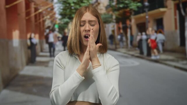 Young blonde woman presses hands to chest while shivering on a city street; vulnerability discomfort resilience.