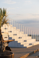 Staircase leading to a seaside restaurant at sunset, with a view of the calm ocean, a sailboat in the distance, and tropical palm leaves in the foreground.
