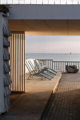 Seaside terrace with empty lounge chairs and a closed umbrella at sunset, overlooking calm ocean water and distant sailboats.