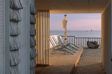 Seaside terrace with empty lounge chairs and a closed umbrella at sunset, overlooking calm ocean water and distant sailboats.
