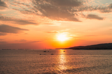 Obraz premium Beautiful sunset over the sea with golden sunlight reflecting on the water, small boats on the horizon, and distant coastline under a dramatic sky.