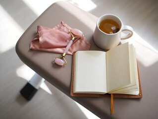 An open journal, a face roller, and a cup of tea are placed on a bench in sunlight