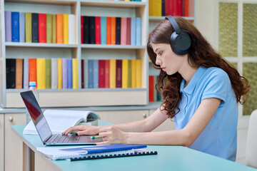 Teenage girl student in headphones studying in educational library, using laptop