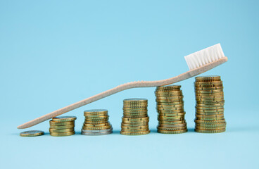 Concept of dental costs increasing. Stacks of coins lined up in ascending order with a beige toothbrush placed diagonally on top of them. Background is light blue. Studio shot.