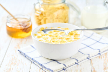 corn flakes in a bowl with milk and honey on a white wooden table.