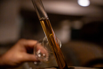 Gros plan sur les mains d’un homme prélevant du champagne avec une pipette dans un tonneau en cave, pour le déguster dans un verre.