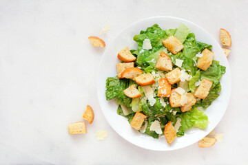 Traditional Caesar salad with croutons and parmesan cheese. Top down view on a white marble background.