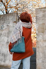 Person with red hair wearing orange coat, gray scarf, and blue bag stands by concrete wall with blooming trees in background