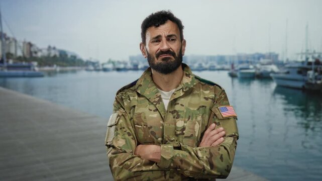 Military man with beard in uniform stands at a bustling port with ships and boats in the background, showcasing a patriotic american flag patch.