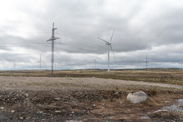 Wind turbines stand tall on a rugged tundra landscape under a dramatic cloudy sky, with muddy...