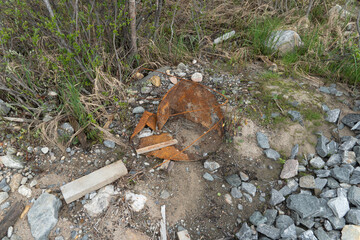 A broken and rusted metal barrel lies abandoned on rocky ground surrounded by wild vegetation and...