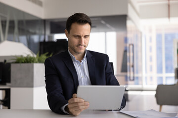 Focused businessman sits alone in modern office, working on digital tablet, look at device screen,...