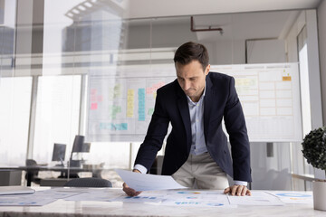 Project manager stand at table holds documents, analyze marketing research results, reviewing...
