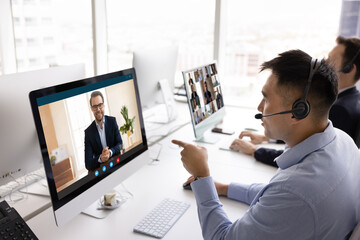 Young 35s Chinese professional man sits at desk in modern coworking office wear headset looks at PC monitor screen displaying young Caucasian businessman engaged in video call, negotiating remotely