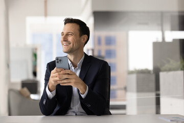 Happy businessman sit in workplace with smartphone, using messaging apps, browsing internet...