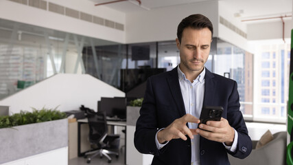 Serious businessman dressed in suit holds smartphone standing alone in contemporary office, sending...