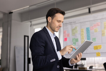 Serious middle-aged businessman dressed in formal suit typing on tablet, stands with gadget in...