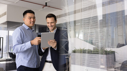 Two smiling men team members standing in office using tablet, editing or reviewing document...