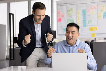 Hispanic and Asian workmates celebrate amazing business news, look at laptop screen, screaming, enjoy promotion, sales or salary growth or professional advancement, reach financial or corporate goal